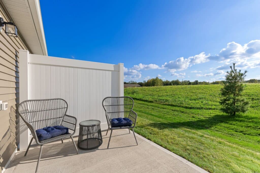 Two black metal chairs with navy cushions and a round glass table sit on a concrete patio overlooking a green grassy field under a blue sky with scattered clouds.