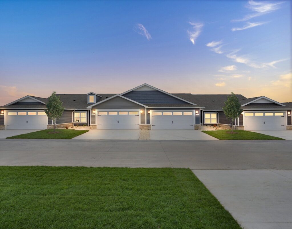 Row of modern duplex homes with attached garages, concrete driveways, small lawns, and a clear sky at sunset.