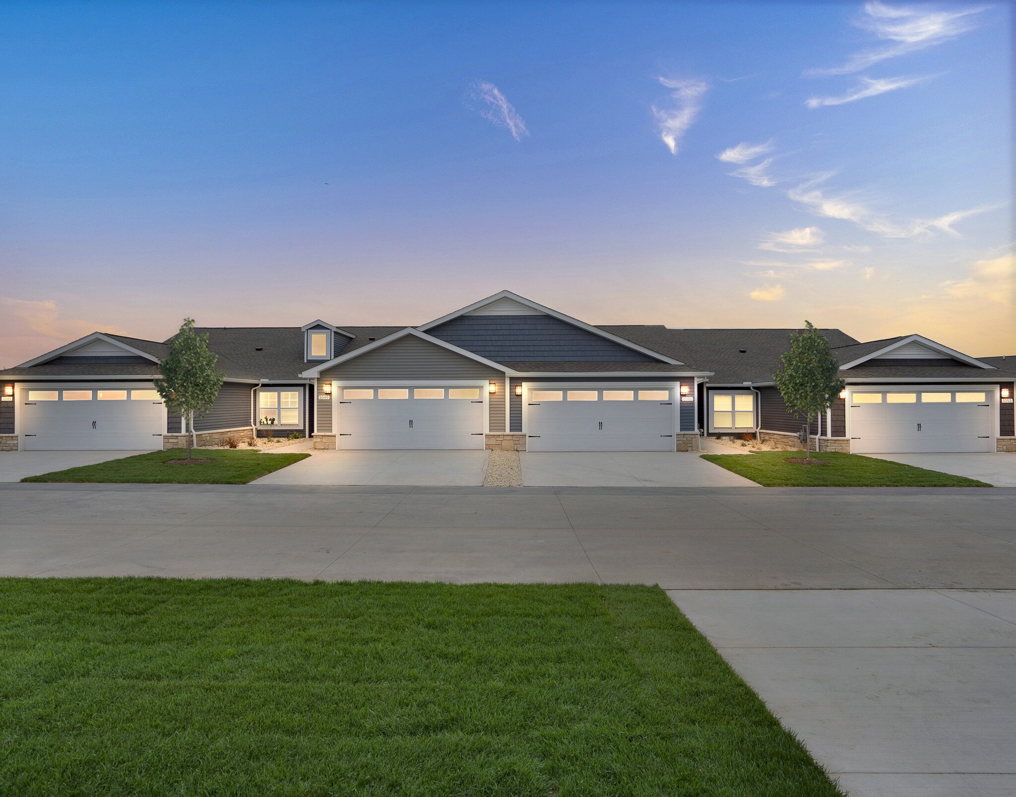 Row of modern duplex homes with attached garages, concrete driveways, small lawns, and a clear sky at sunset.