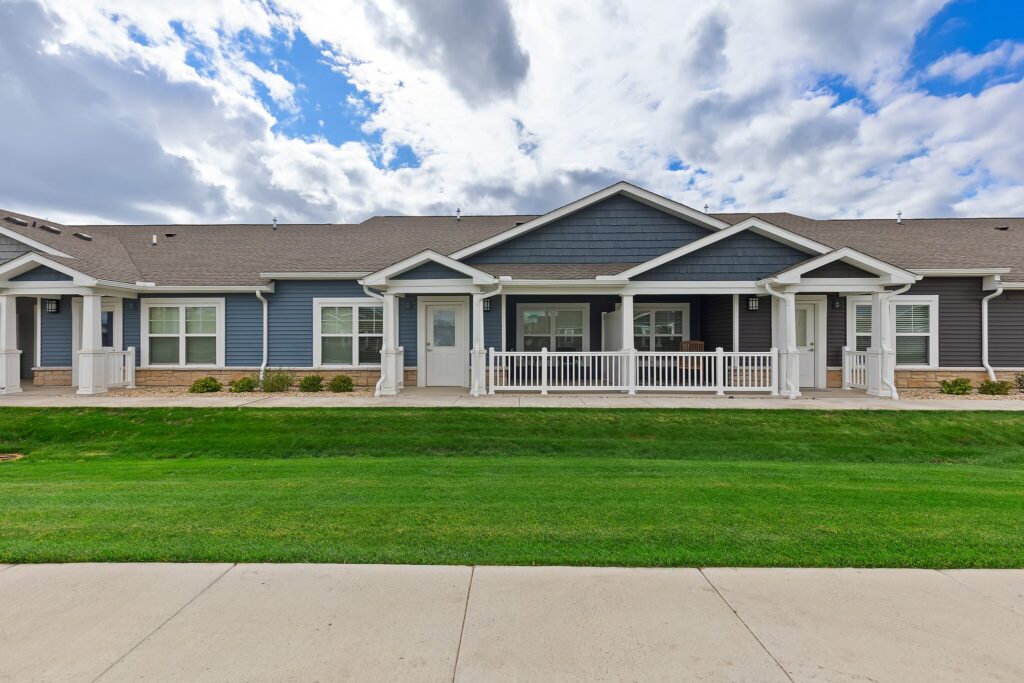 Single-story residential building with a covered front porch, white railings, blue and gray siding, and a well-maintained lawn under a partly cloudy sky.
