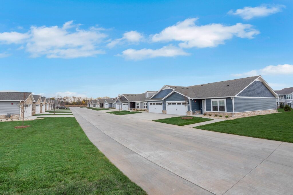 A suburban street with single-story houses, green lawns, and a clear blue sky.