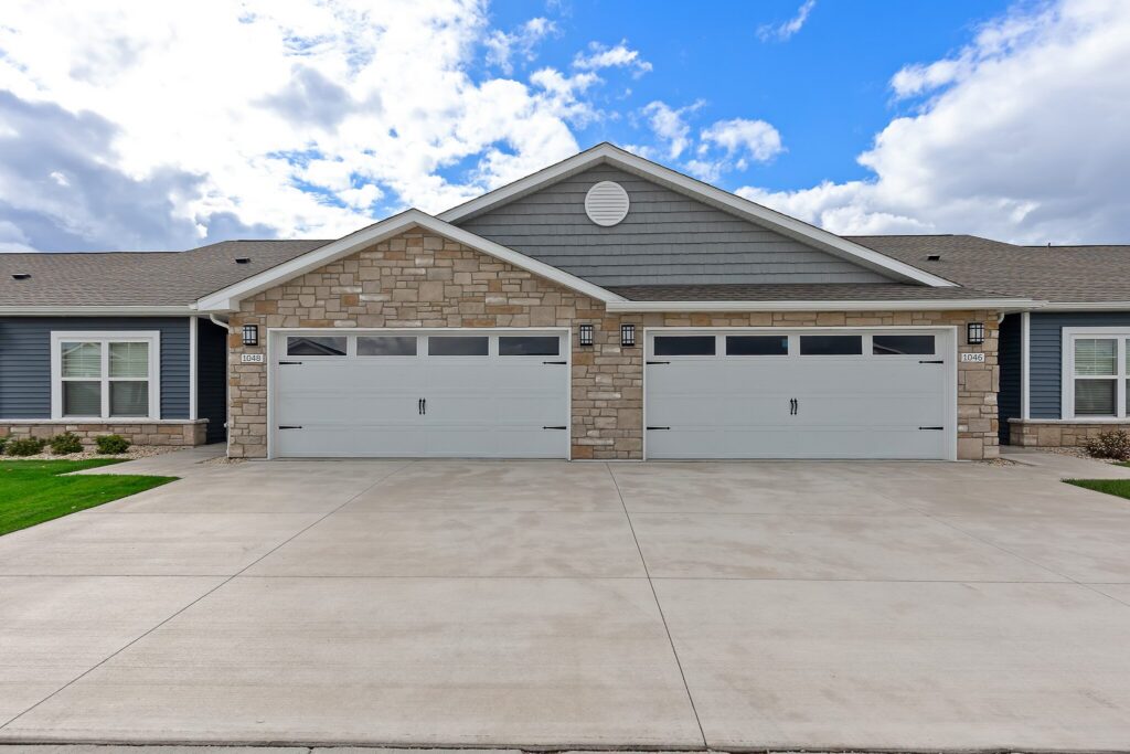 A duplex house with stone and gray siding exterior, each unit featuring a double garage and a wide concrete driveway under a partly cloudy sky.