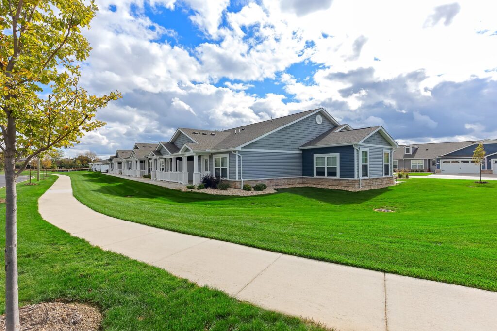 Single-story blue and white houses with manicured lawns line a curved sidewalk under a partly cloudy sky.