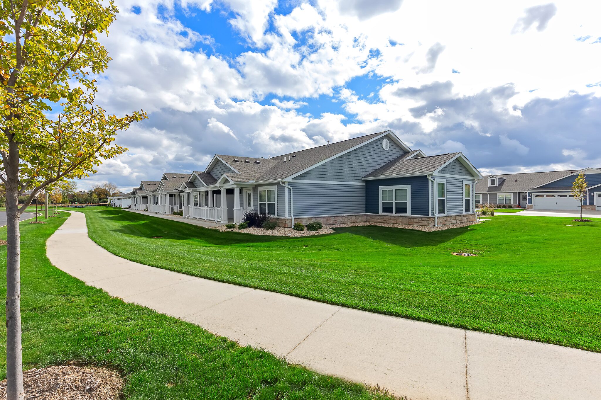 Single-story blue and white houses with manicured lawns line a curved sidewalk under a partly cloudy sky.