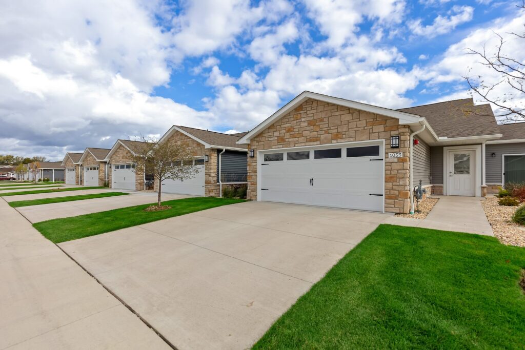 Row of modern single-story townhouses with stone facades, white garage doors, and neat green lawns under a partly cloudy sky.