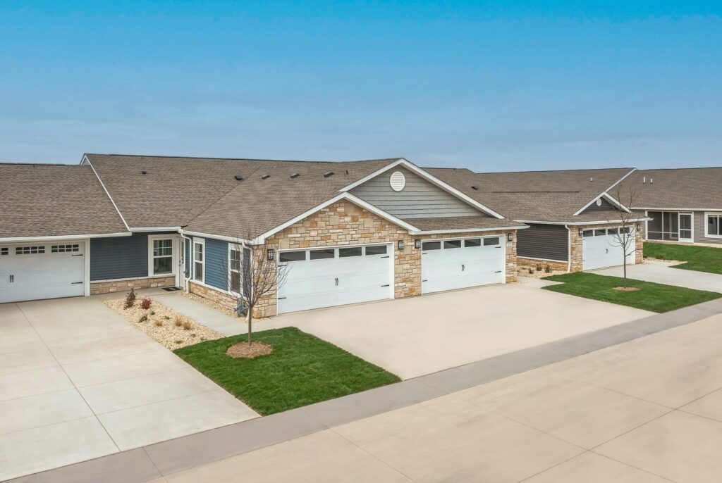 Row of single-story townhomes with attached two-car garages, stone and siding exteriors, manicured lawns, and concrete driveways under a clear blue sky.