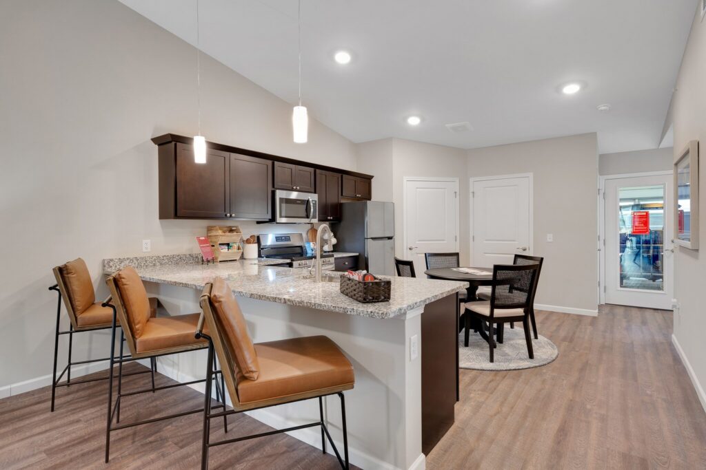 Modern kitchen with granite countertops, brown cabinets, stainless steel appliances, a breakfast bar with three chairs, and a small dining area with a round table and four chairs.