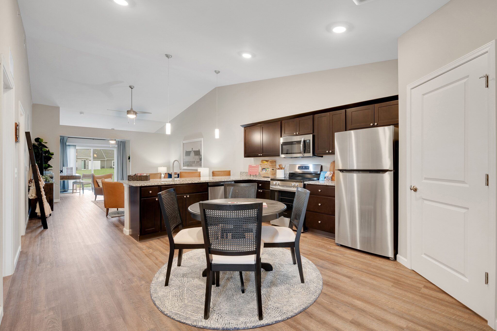 Modern kitchen and dining area with dark wood cabinets, stainless steel appliances, round table with four chairs, and wood flooring; living room visible in the background.