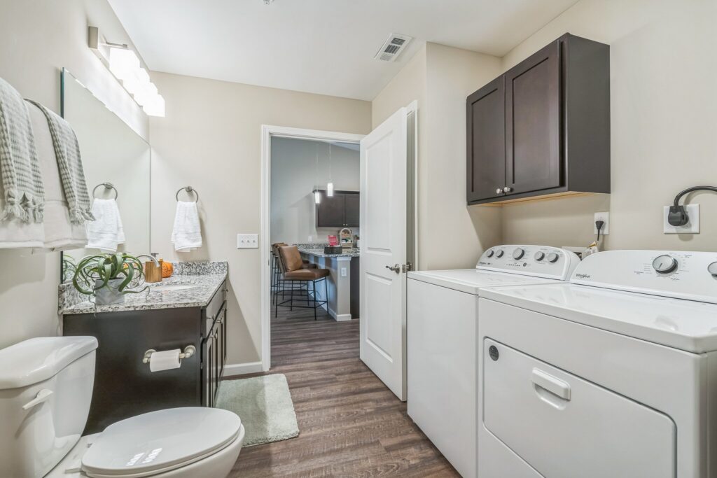 Bathroom and laundry room combination with a toilet, sink, cabinets, washer, and dryer; view into a kitchen area through an open door.