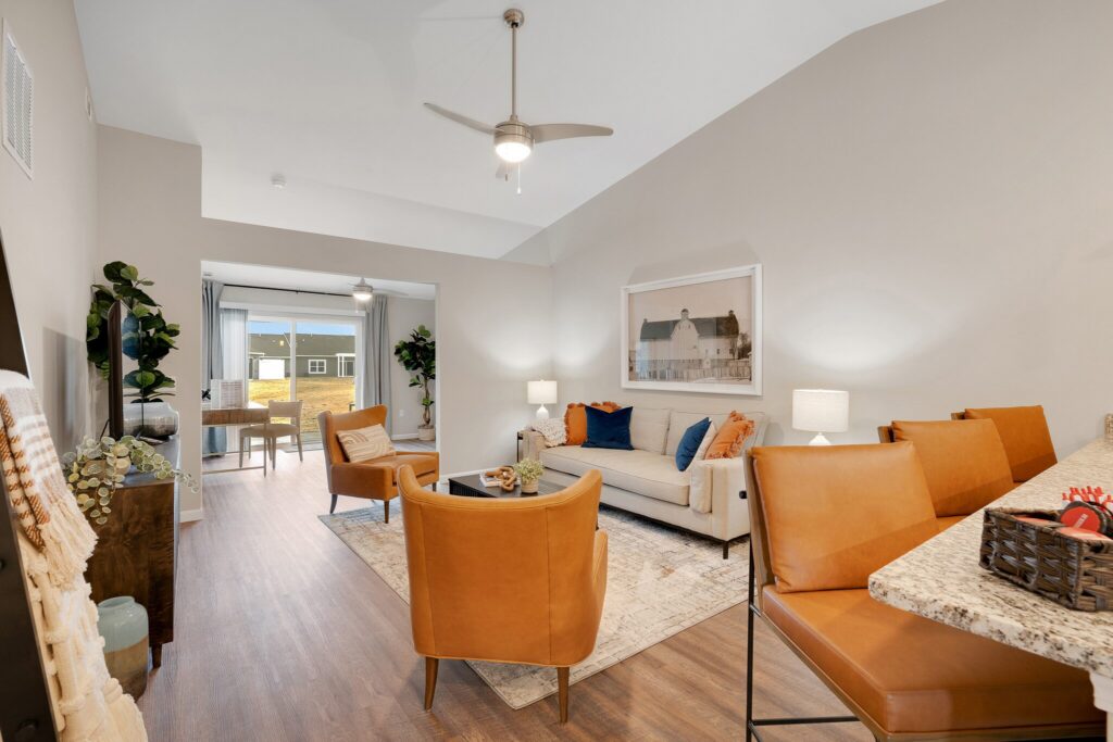 Modern living room with tan and orange furniture, a ceiling fan, wooden floors, and a small dining area with granite countertop, well-lit by natural and artificial light.