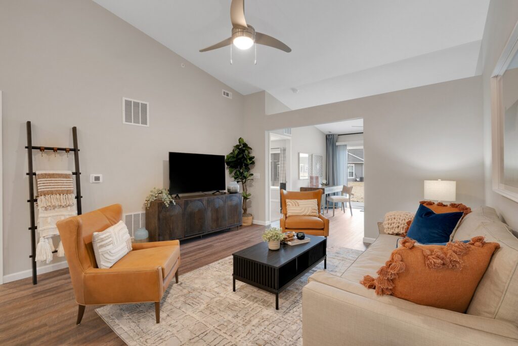 Modern living room with beige walls, a cream sofa with orange pillows, two orange armchairs, a TV on a dark cabinet, a coffee table, and a ceiling fan. Dining area visible in the background.
