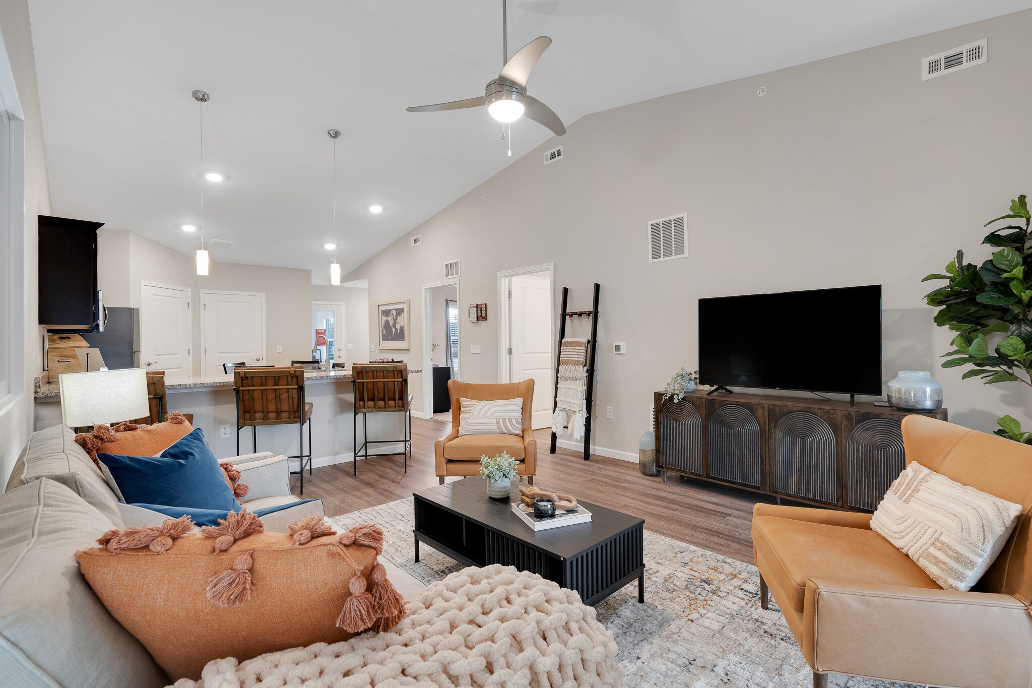 Modern living room with beige walls, a sofa, two orange armchairs, a coffee table, TV on a cabinet, bar stools at a kitchen counter, and decorative plants.