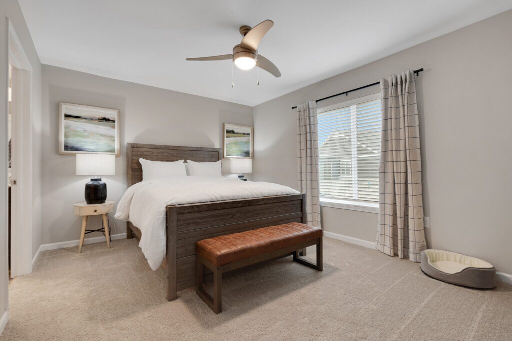 A bedroom with a wooden bed, white bedding, two bedside tables with lamps, a bench at the foot of the bed, a window with curtains, and a ceiling fan.