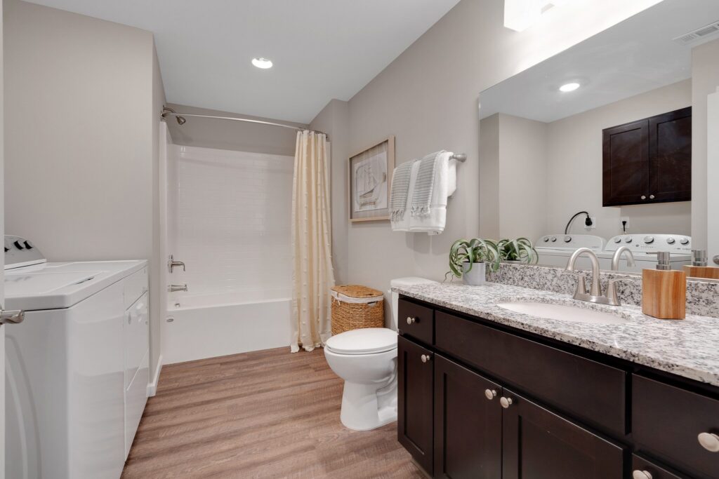 Modern bathroom with laundry machines, a bathtub with curtain, toilet, dark wood vanity with granite countertop, large mirror, and towels hanging on the wall.