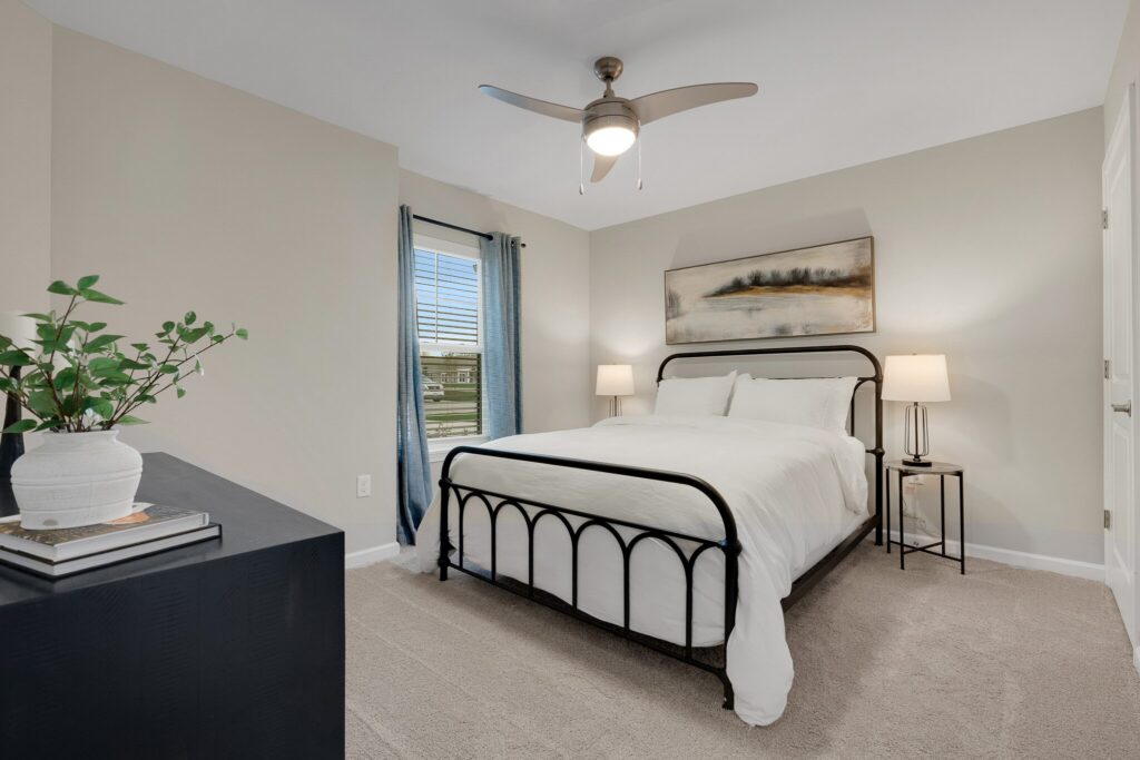 A modern bedroom with a black metal bed frame, white bedding, two bedside tables with lamps, a potted plant on a dresser, and a landscape painting above the bed.