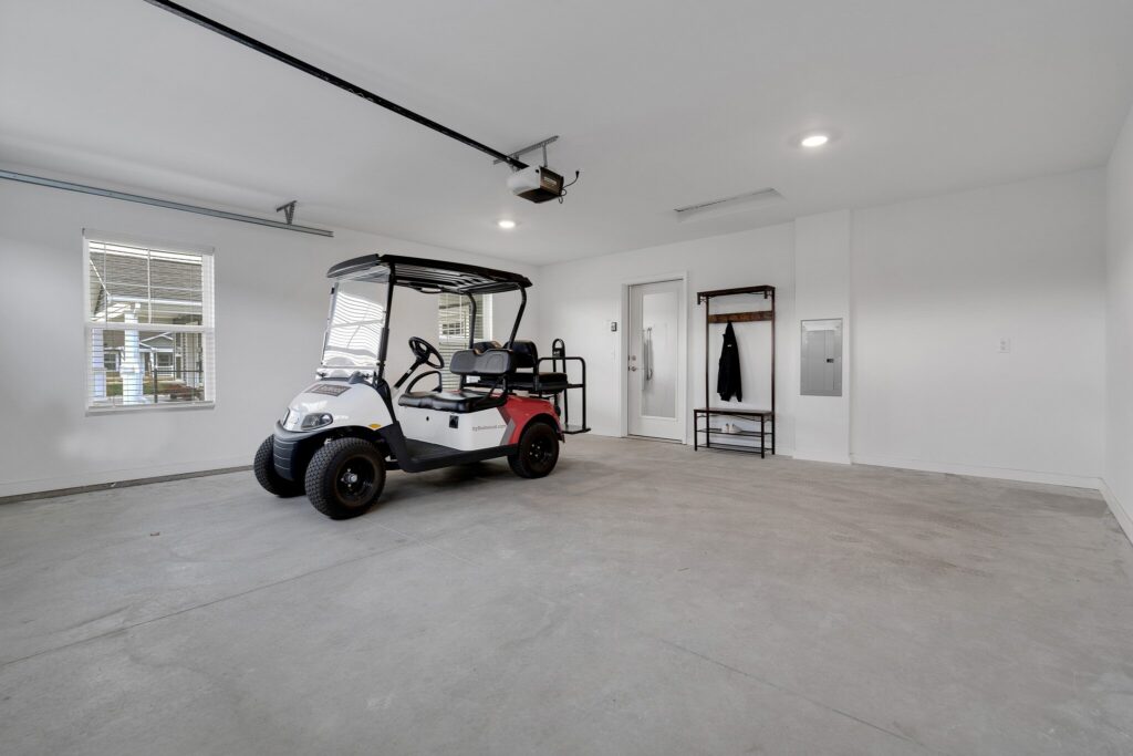 A golf cart is parked inside a clean, spacious garage with white walls, two windows, and a small coat rack by the door.