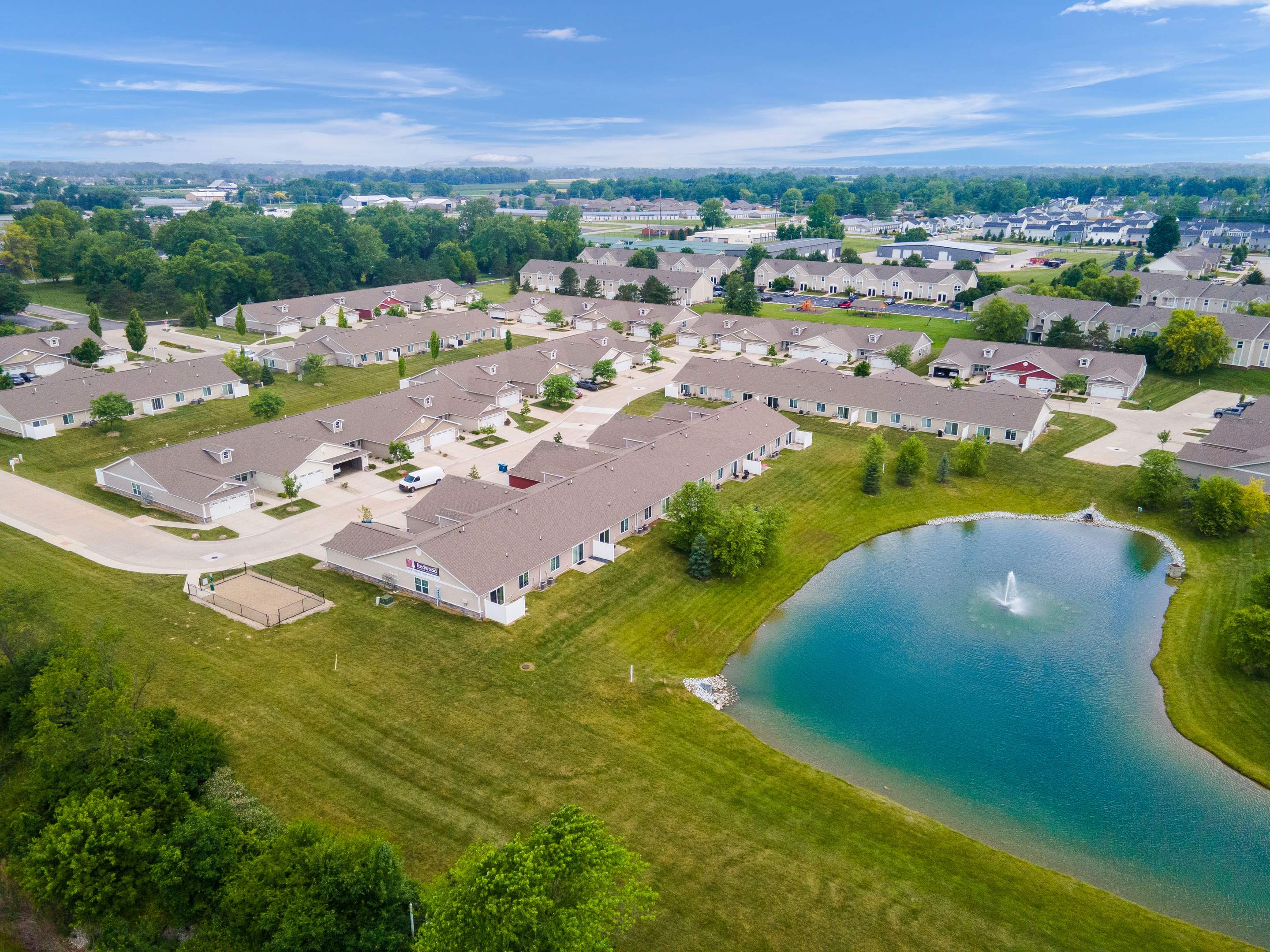 Aerial view of a suburban neighborhood with single-story houses, a small pond with a fountain, and green lawns under a partly cloudy sky.