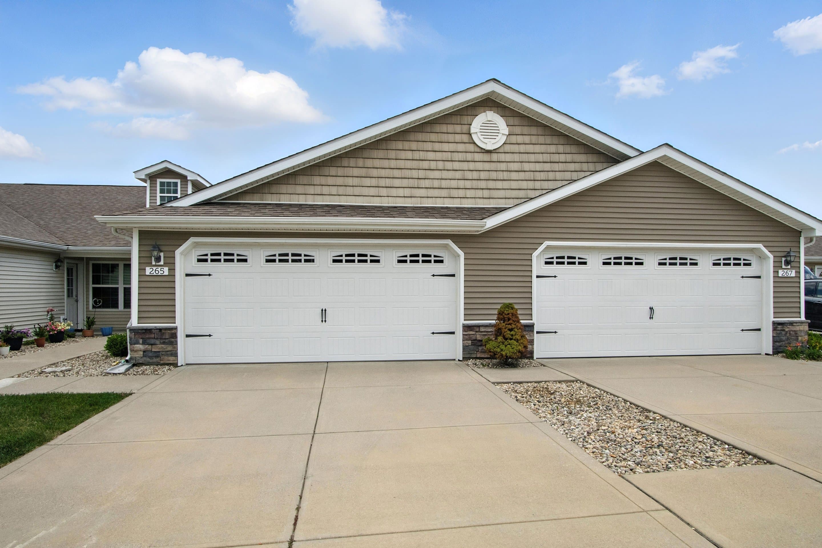 Duplex house with two attached double-car garages, beige siding, stone accents, concrete driveway, and minimal landscaping under a blue sky with clouds.