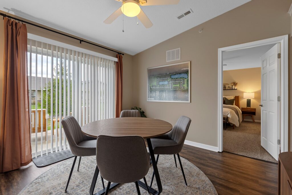 A dining area with a round table and four chairs, vertical blinds on a sliding door, and an open doorway leading to a bedroom.