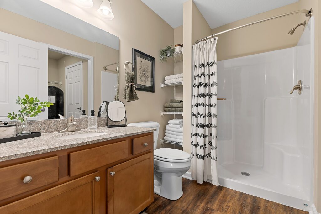 A bathroom with a wooden vanity, granite countertop, toilet, open shelving with towels, and a walk-in shower with a white curtain and metal fixtures.