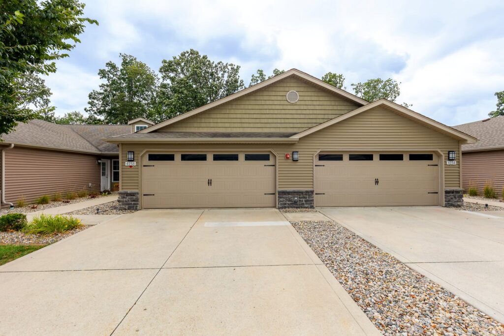 A beige duplex with two double garages, stone accents, and a concrete driveway, surrounded by minimal landscaping and decorative rocks.