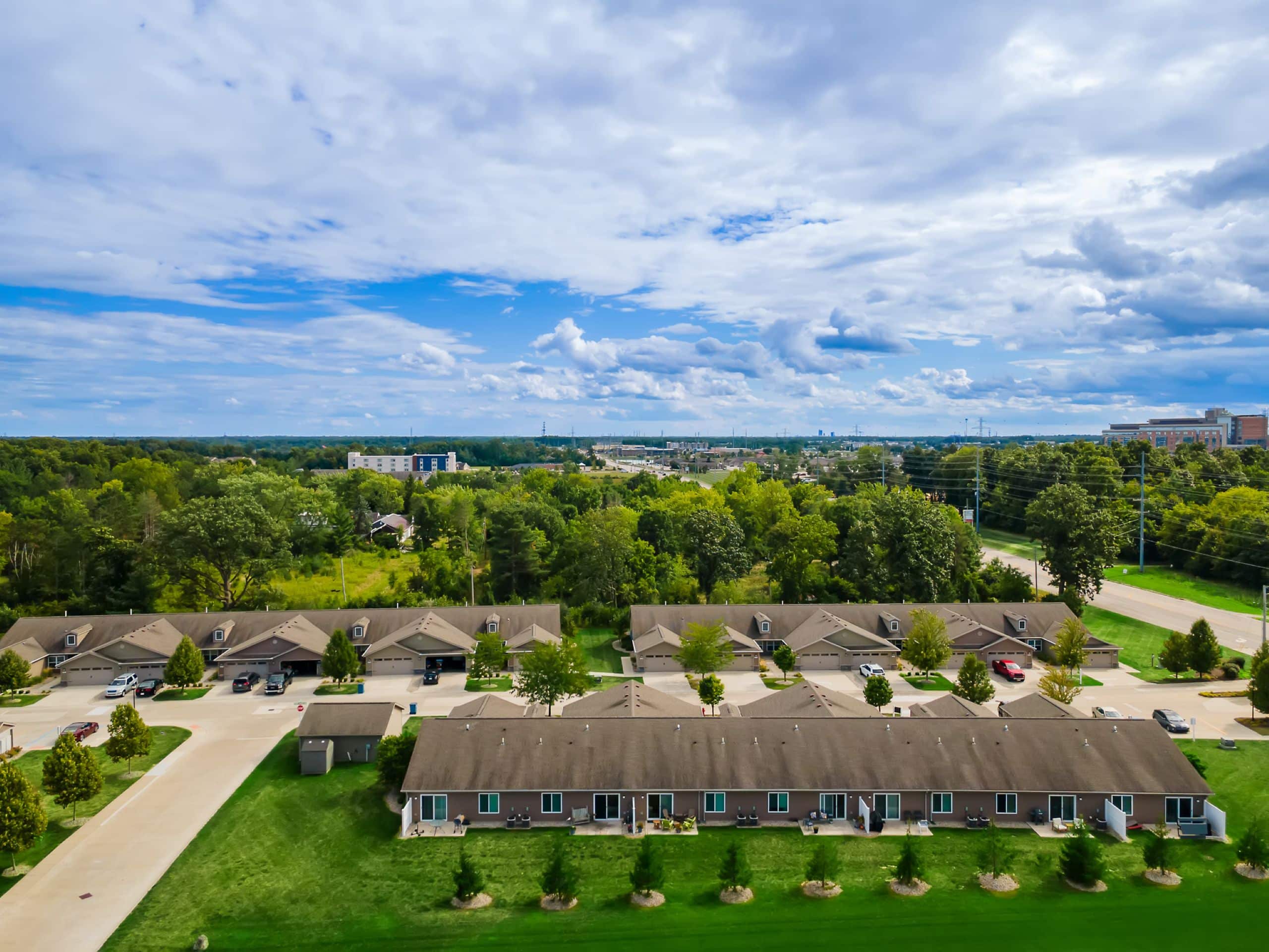 Aerial view of a row of single-story townhouses with driveways, a green lawn, trees, and a road with cars; buildings and forested area in the background under a cloudy sky.