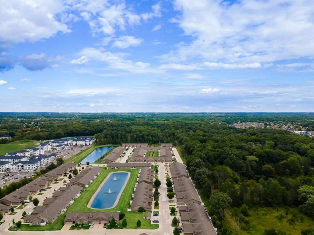 Aerial view of a suburban neighborhood with rows of houses, landscaped ponds with fountains, and a large expanse of trees under a partly cloudy sky.