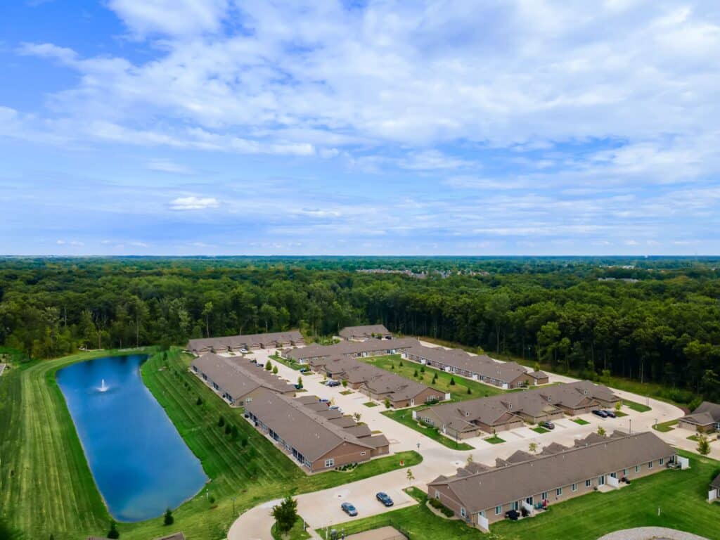 Aerial view of a suburban residential complex with single-story buildings, a pond with a fountain, and surrounding trees under a partly cloudy sky.