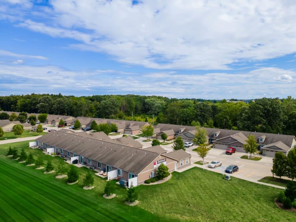 Aerial view of a residential neighborhood with single-story homes, green lawns, neatly trimmed bushes, parked cars, and trees in the background under a partly cloudy sky.