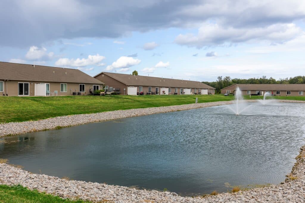 Single-story apartment buildings stand behind a small pond with fountains, surrounded by grass and rocks under a partly cloudy sky.