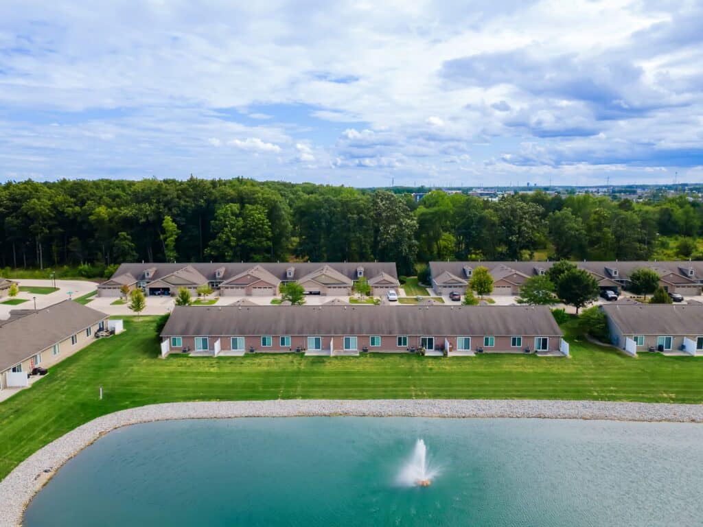 Aerial view of single-story residential buildings with a pond and fountain in the foreground, bordered by green lawns and trees under a partly cloudy sky.