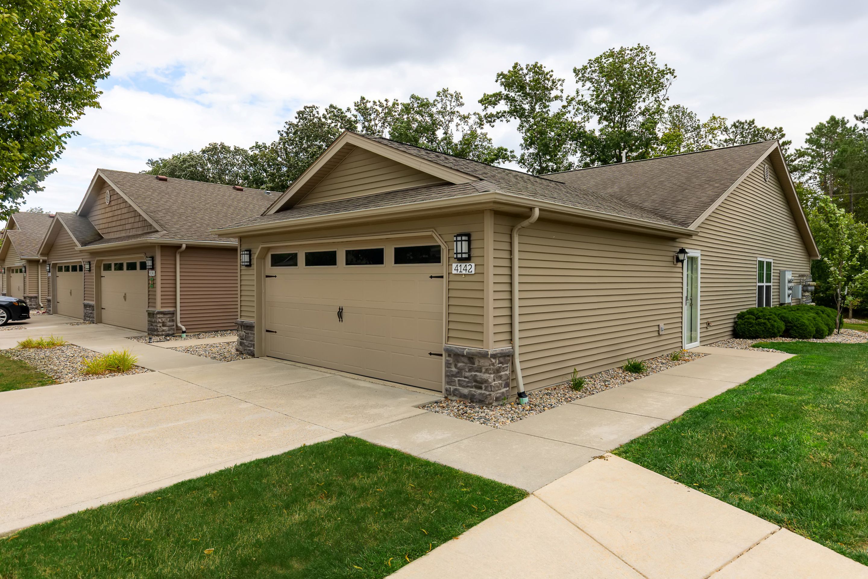 Single-story suburban house with beige siding, a double garage, stone accents, and a concrete driveway, surrounded by grass and small shrubs.