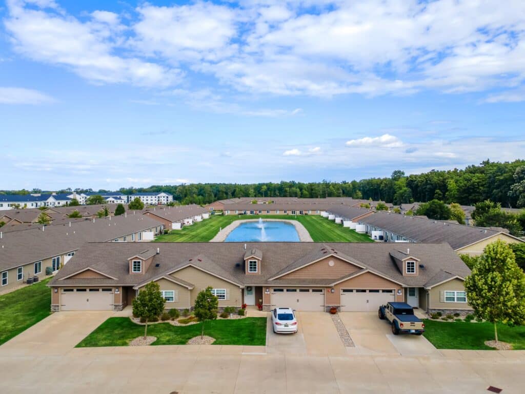 Aerial view of a suburban neighborhood featuring single-story homes, a central pond, green lawns, and parked cars under a partly cloudy sky.