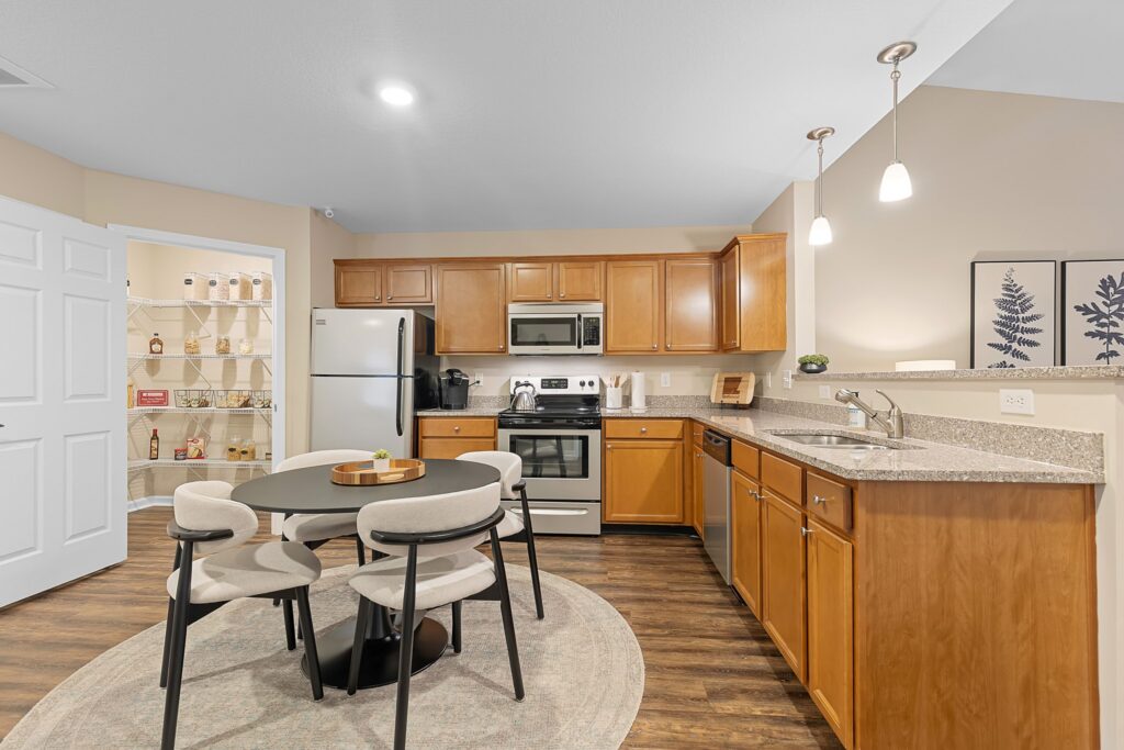 Modern kitchen with stainless steel appliances, wooden cabinets, round dining table with four chairs, and an open pantry stocked with various items.