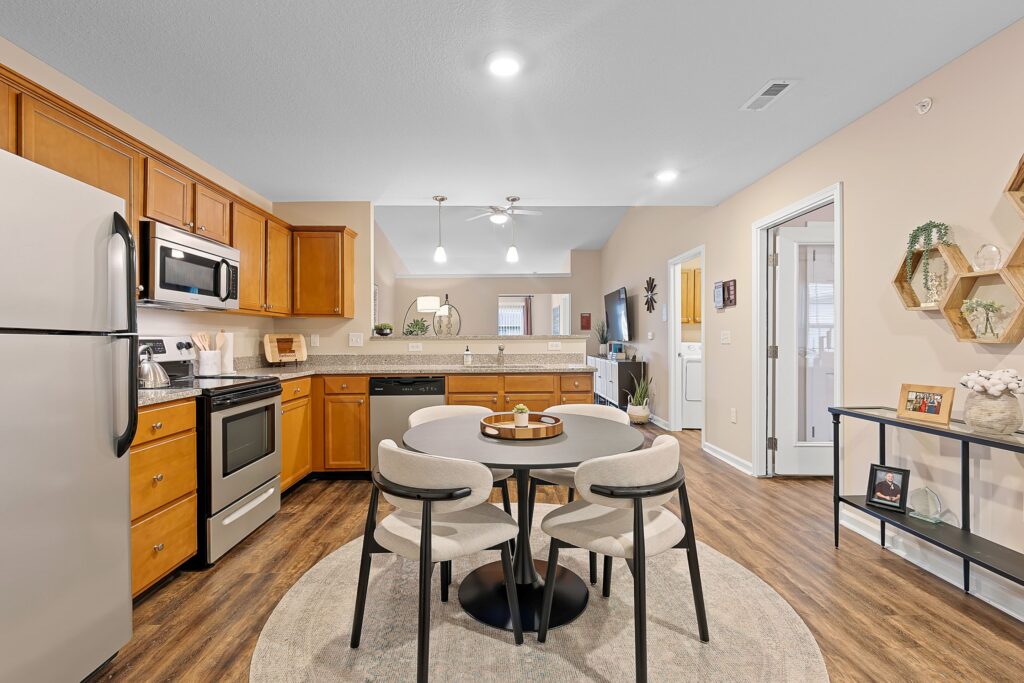 Modern kitchen and dining area with stainless steel appliances, wooden cabinets, a round dining table with four chairs, and neutral-colored decor on hardwood flooring.