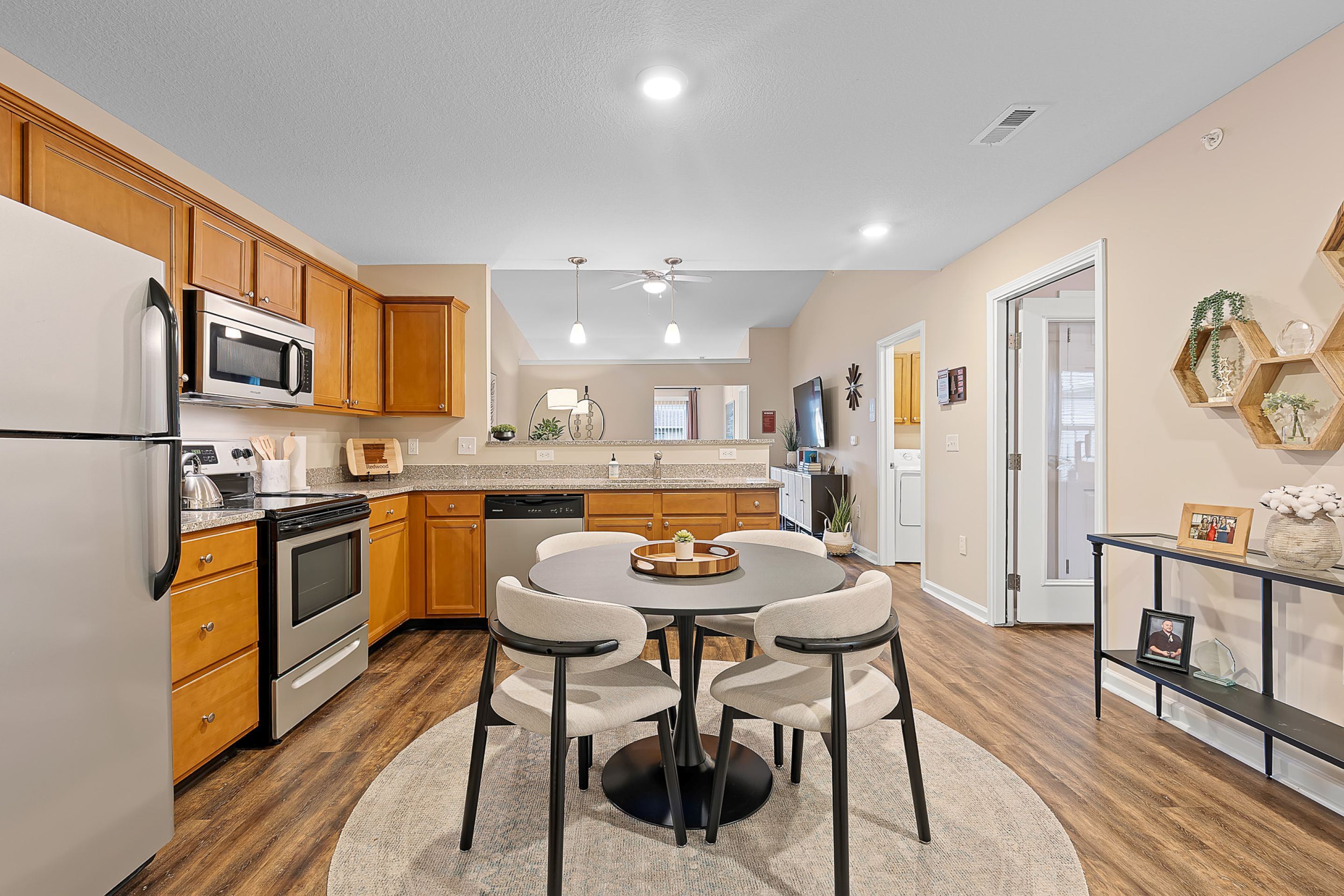 Modern kitchen and dining area with stainless steel appliances, wooden cabinets, a round dining table with four chairs, and neutral-colored decor on hardwood flooring.