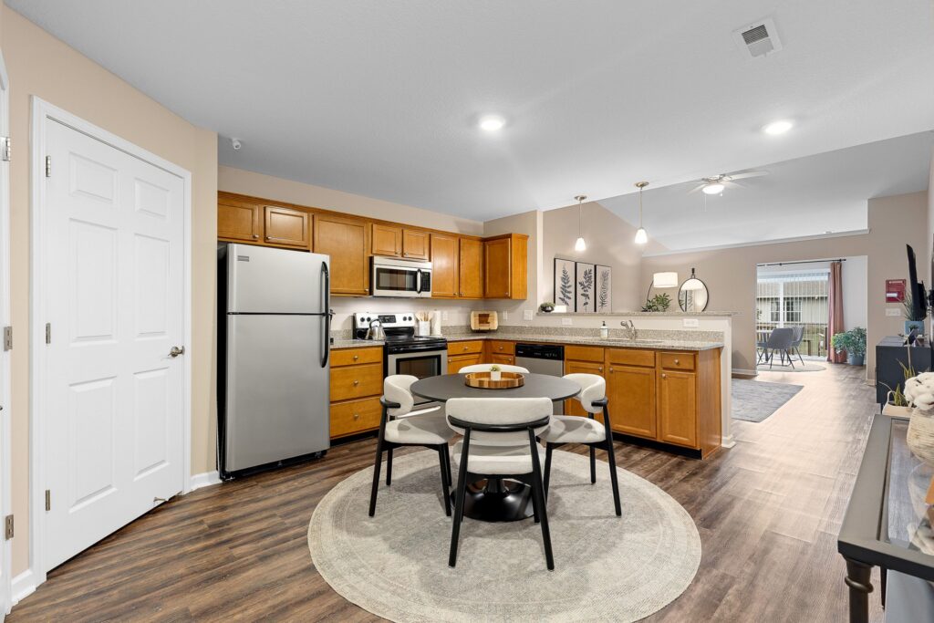 Modern kitchen and dining area with stainless steel appliances, wooden cabinets, round table with four chairs, and hardwood floors, open to a living space in the background.