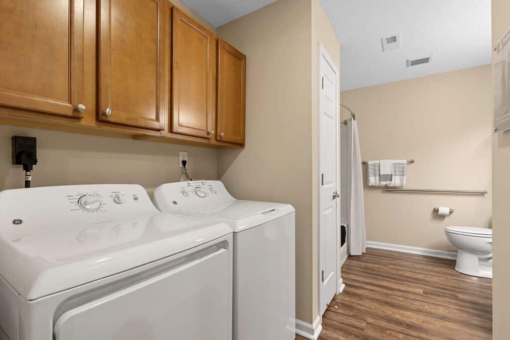 Laundry room with a white washer and dryer, wooden cabinets above, beige walls, wood flooring, and a bathroom area with a toilet and towel rack in the background.