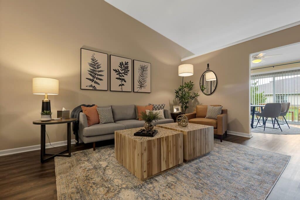 A modern living room with a gray sofa, brown armchair, two wooden coffee tables, botanical artwork, and neutral decor, leading to a sunroom with large windows.