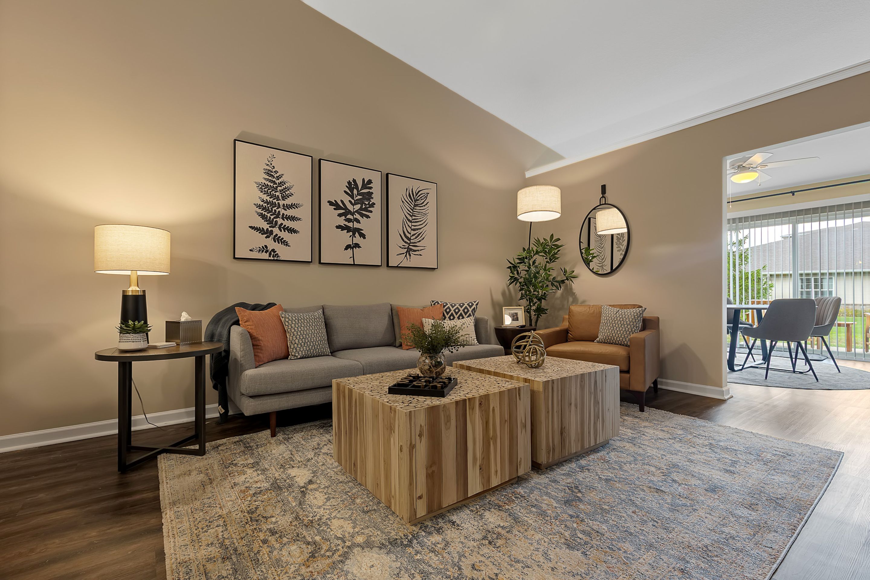 A modern living room with a gray sofa, brown armchair, two wooden coffee tables, botanical artwork, and neutral decor, leading to a sunroom with large windows.
