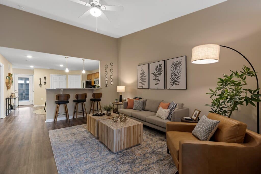 Modern open-concept living room and kitchen with earth-tone decor, a gray sofa, tan armchair, barstools at the counter, and botanical artwork on the wall.