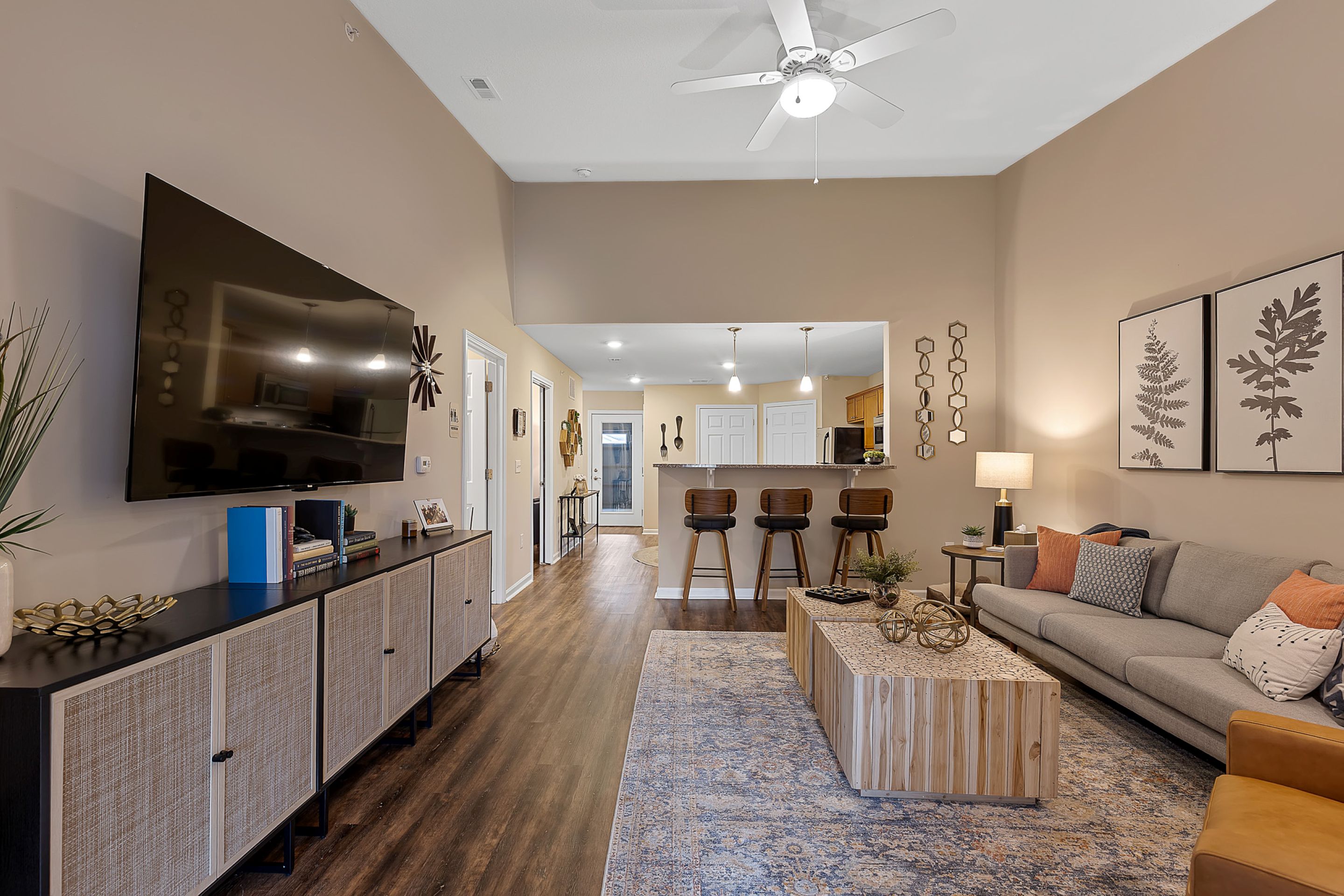 Modern living room with wood flooring, a large wall-mounted TV, a gray sofa, a patterned rug, neutral décor, and a kitchen with bar stools visible in the background.