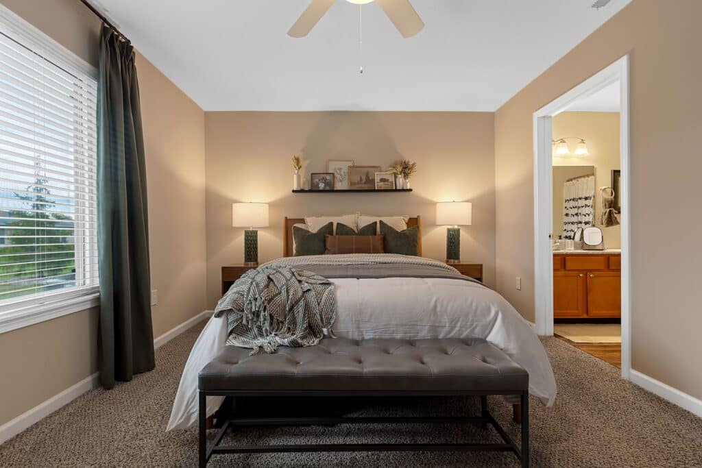 A neatly made bed with a gray bench at the foot, flanked by two lamps, in a beige bedroom with an open doorway leading to a bathroom.