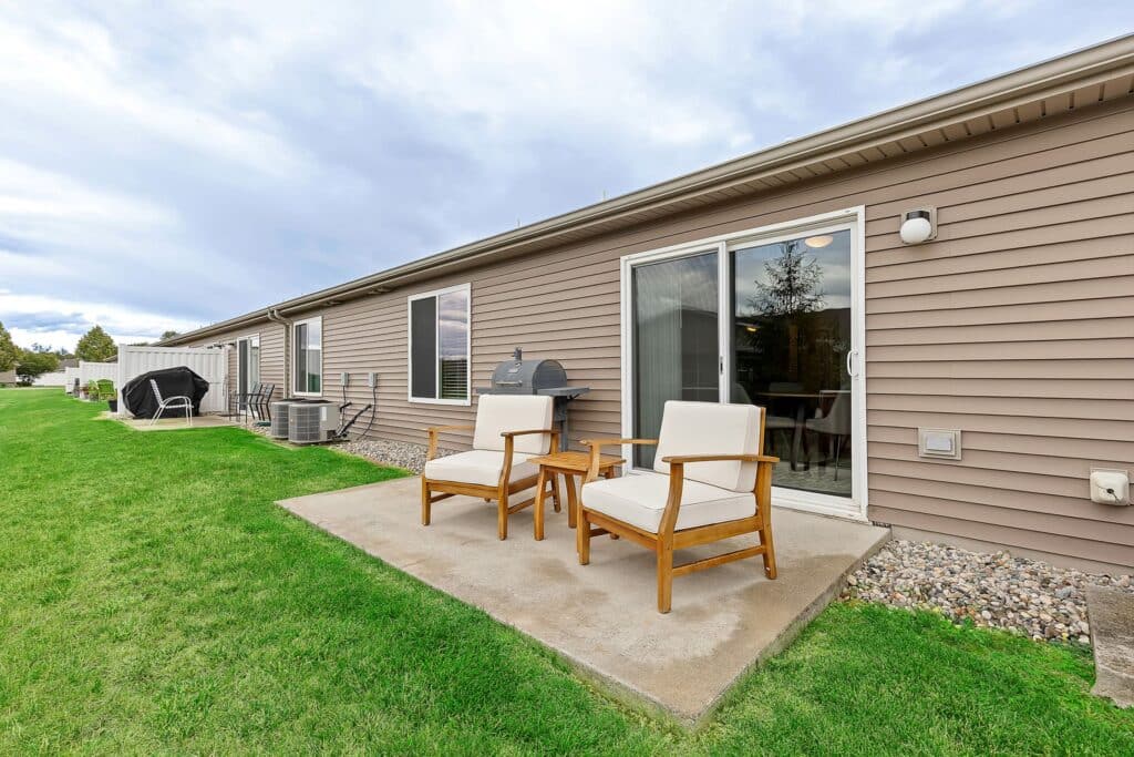 Two cushioned chairs and a small table on a concrete patio outside a beige house with sliding glass doors and a grill, surrounded by green grass.