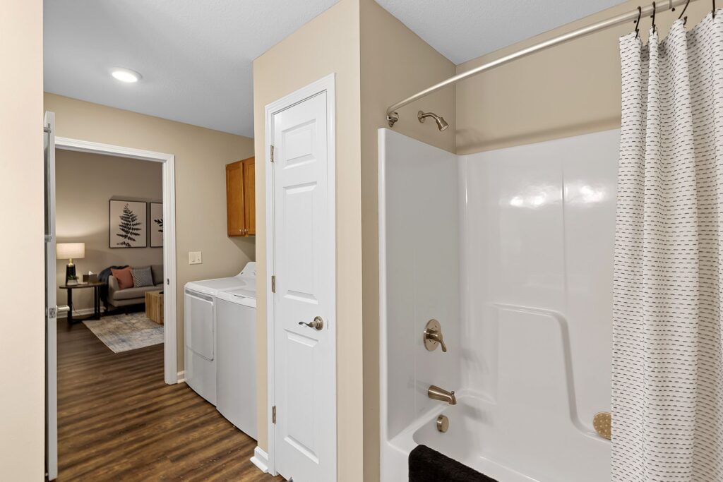 Bathroom with a white bathtub and shower curtain, adjacent to a laundry area with a washer and dryer; living room visible in the background.