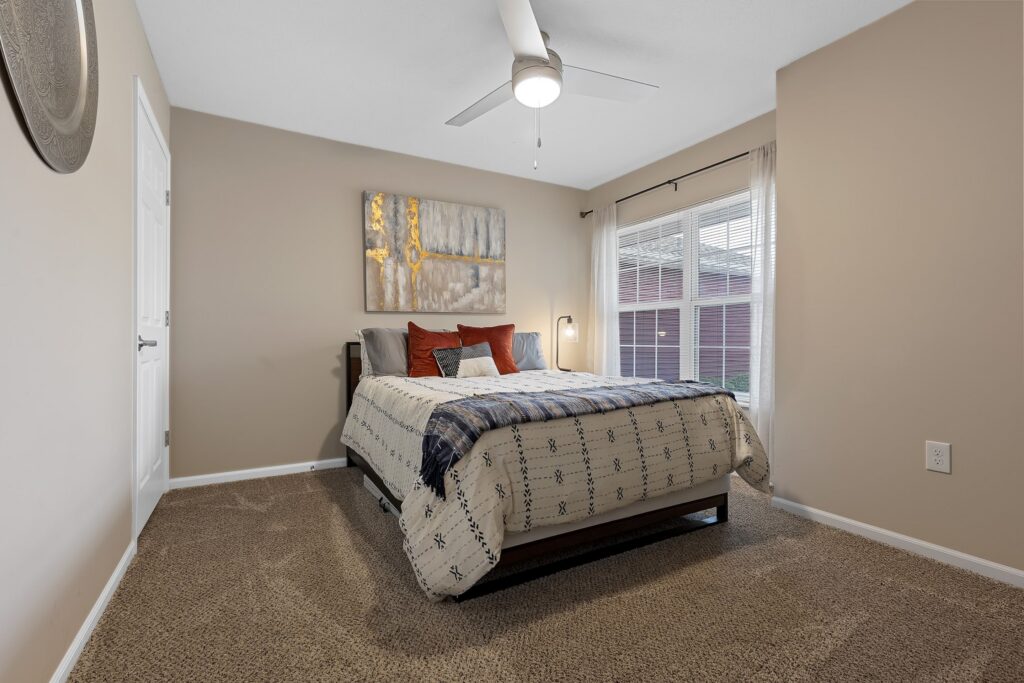 A neatly made bed with patterned bedding and pillows sits in a carpeted bedroom with beige walls, a ceiling fan, a window with blinds, and a piece of artwork above the bed.