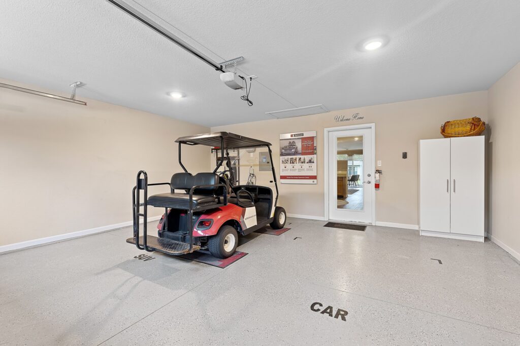 A clean garage interior with a red golf cart parked in a designated spot, white storage cabinet, wall decor, and visible doorway to another room.