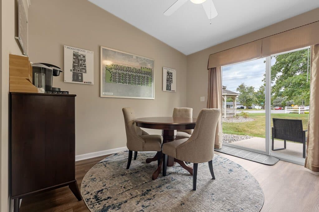 A small dining area with a round table and three chairs on a patterned rug, next to a sliding glass door with a view of a patio and greenery outside.