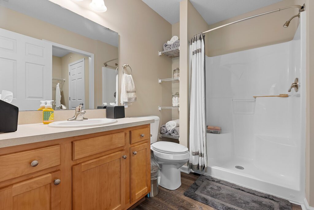 A bathroom with a wooden vanity, sink, mirror, toilet, shelves with towels, and a walk-in shower with a curtain.