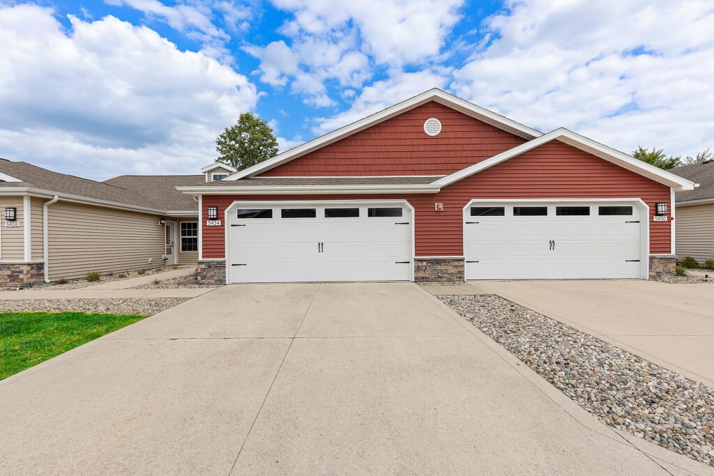 Two attached suburban houses with red and beige siding, each with a double garage and concrete driveway under a partly cloudy sky.
